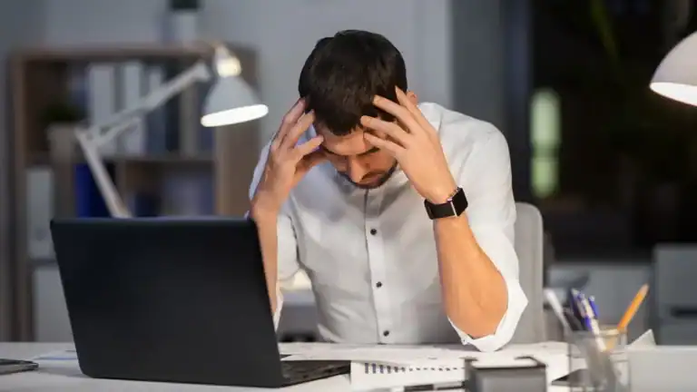 Frustrated man holding head at desk with laptop and office background.